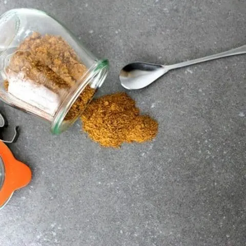 overhead of spicy Chai Spice Mix spilling out of a glass jar on to a table with a spoon next to it.
