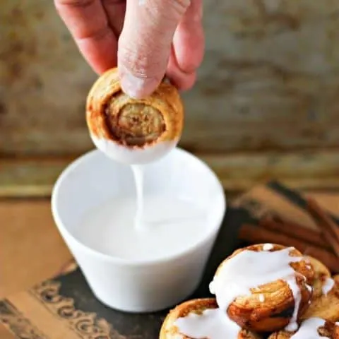 Hand dipping a cinnamon roll in to a small white bowl of icing
