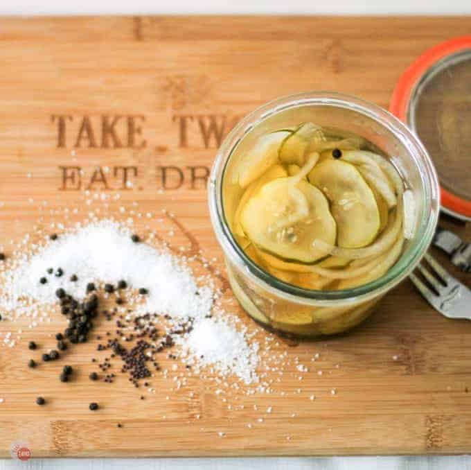 Overhead of homemade pickles in a glass jar with pickling spice beside it on a cutting board