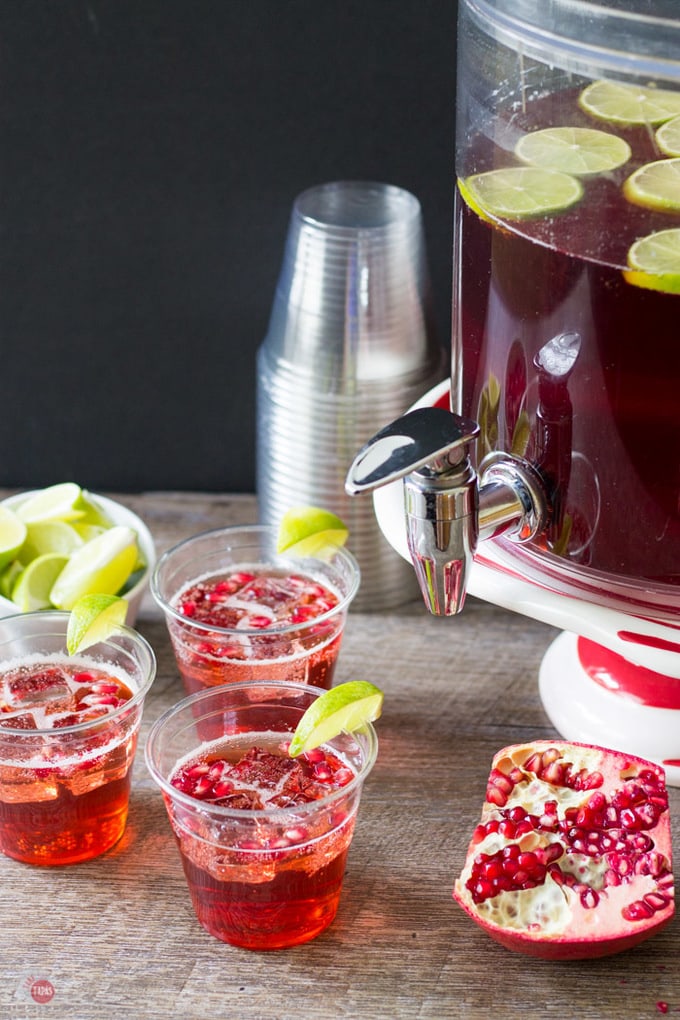 A canister filled with punch, plastic cups, bowl of quartered limes and several glasses filled with the punch.