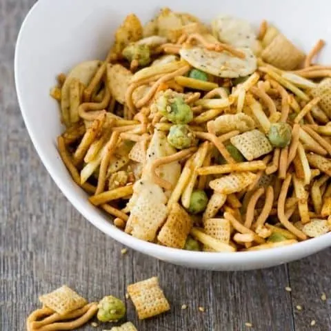 Snack mix in a white bowl with some spilled out on to a wood surface.
