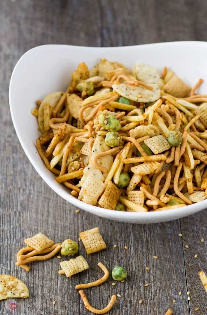 Snack mix in a white bowl with some spilled out on to a wood surface.