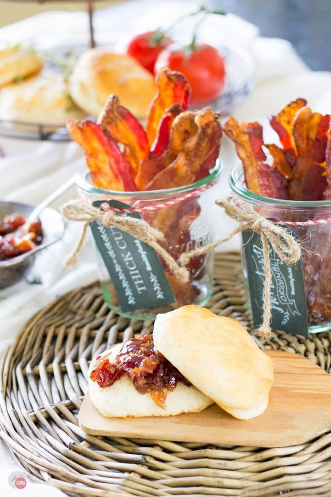 A biscuit with bacon jam on a wood plate and 2 glass jars with bacon in the all on a set table.