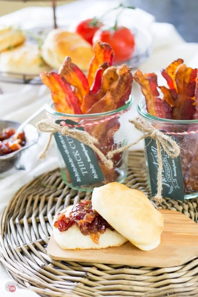 A biscuit with bacon jam on a wood plate and 2 glass jars with bacon in the all on a set table.