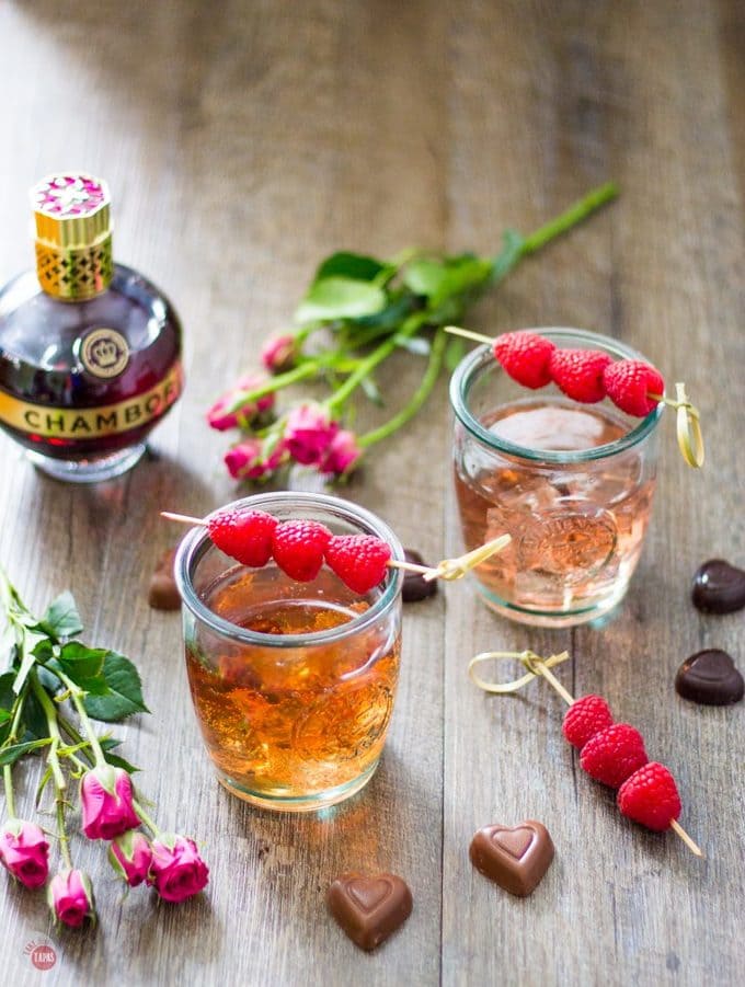 overhead of Chambord Raspberry liqueur, 2 raspberry love cocktails, chocolates and flowers on a wood surface