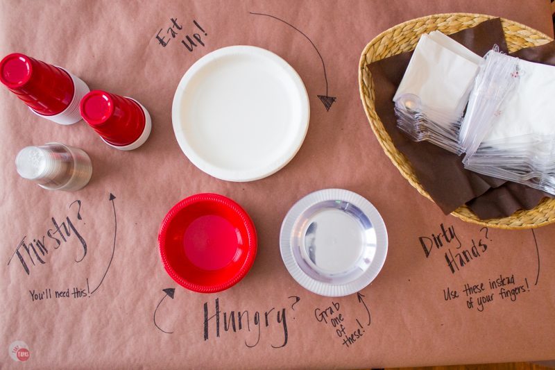 clear and red plastic cups, white plastic plates,red and clear plastic bowls and napkins in a basket all on a brown paper surface