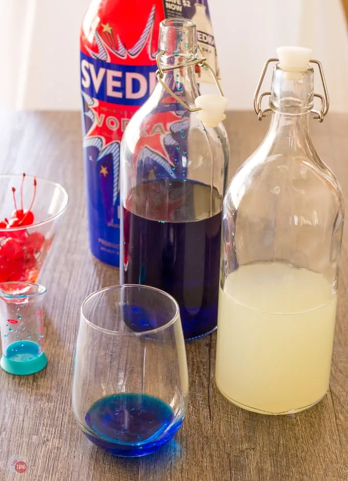 A cocktail glass with blue curacao in it, a bottle of blue liquor, a bottle of white limeade and a bottle of vodka on a wood table