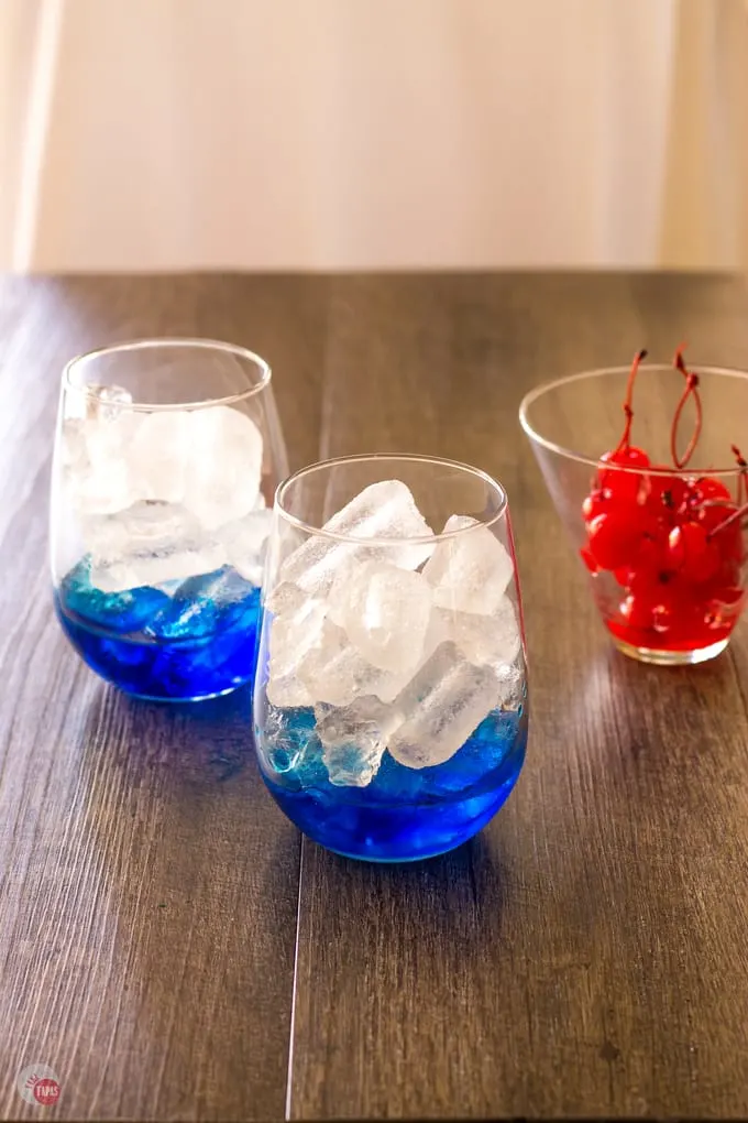 blue curacao in the bottom of 2 cocktail glasses and a small cup of maraschino cherries on a wood surface