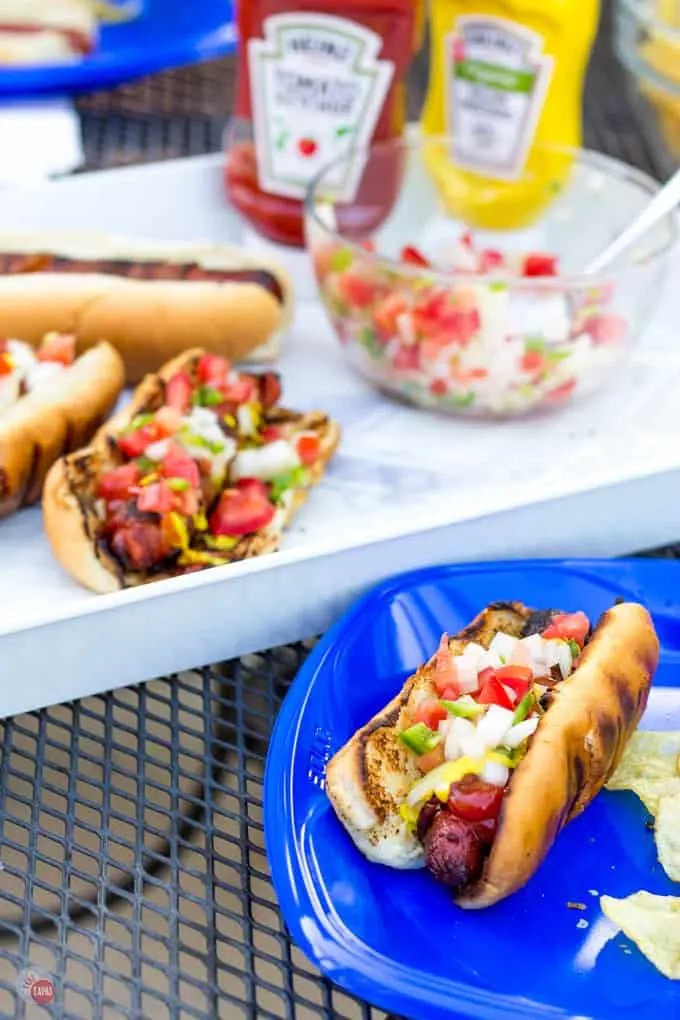 Bacon Wrapped Hot Dog loaded up with jalapeno relish on a blue plate. Other hot dogs on a white platter in the background.