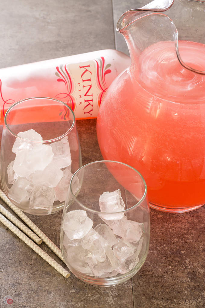 2 glasses with ice, straws, a bottle of pink liquor and a glass pitcher ful of Pink passion party punch on a gray surface