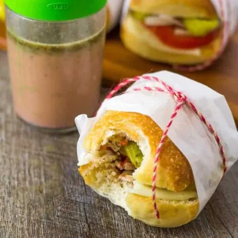 A Mini Italian Veggie Slider Sandwich with a bite taken out of it, sitting on a wood table with dressing and another slider in the background.