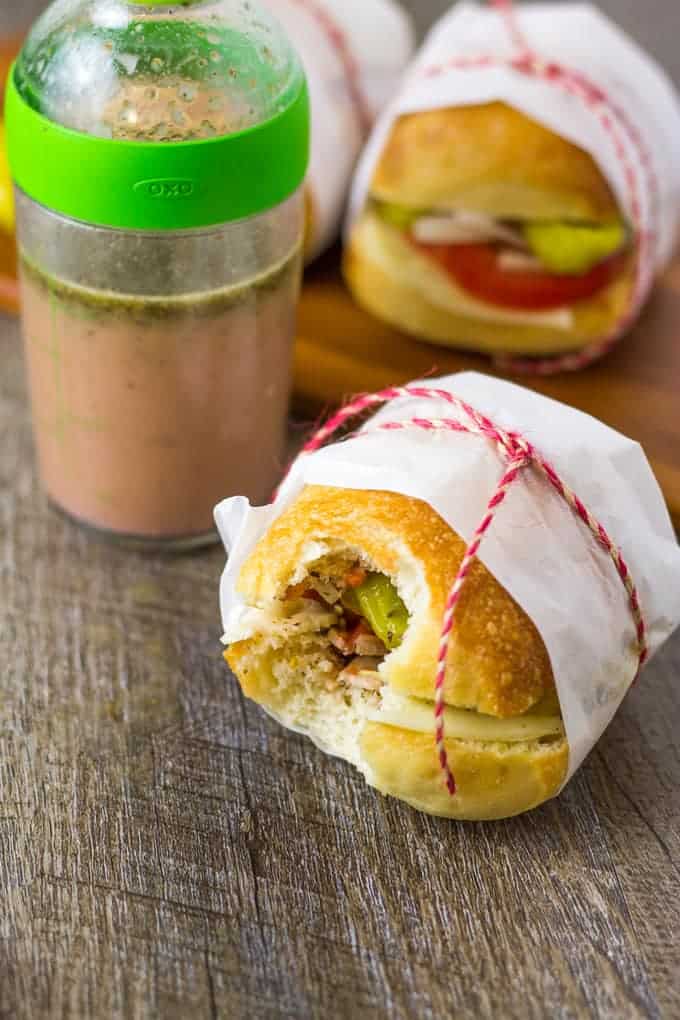 A Mini Italian Veggie Slider Sandwich with a bite taken out of it, sitting on a wood table with dressing and another slider in the background.