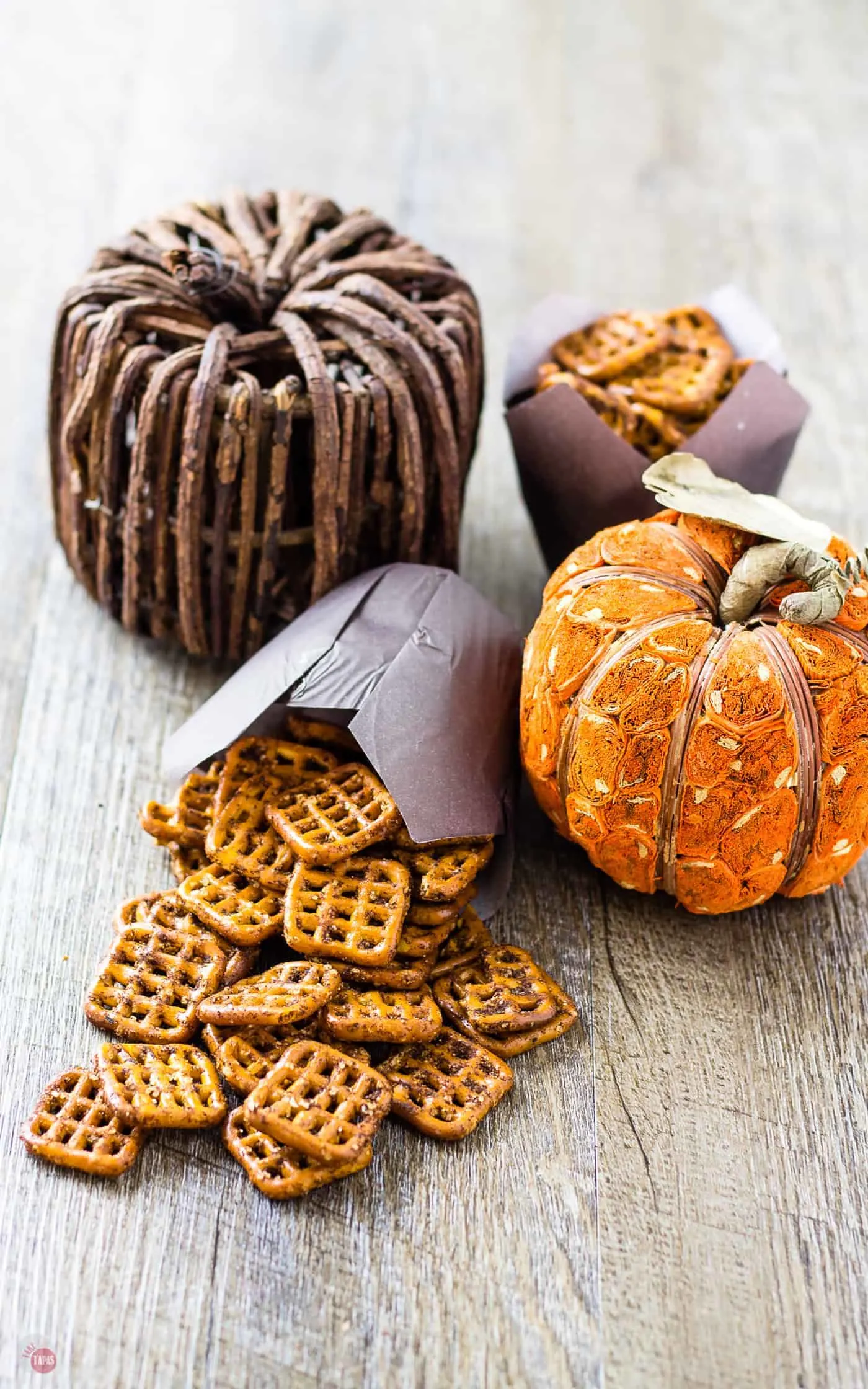 Pumpkin Spice Espresso Pretzels spilling out of a cupcake holder, a full cupcake wrapper and 2 pumpkin decorations on a wood table