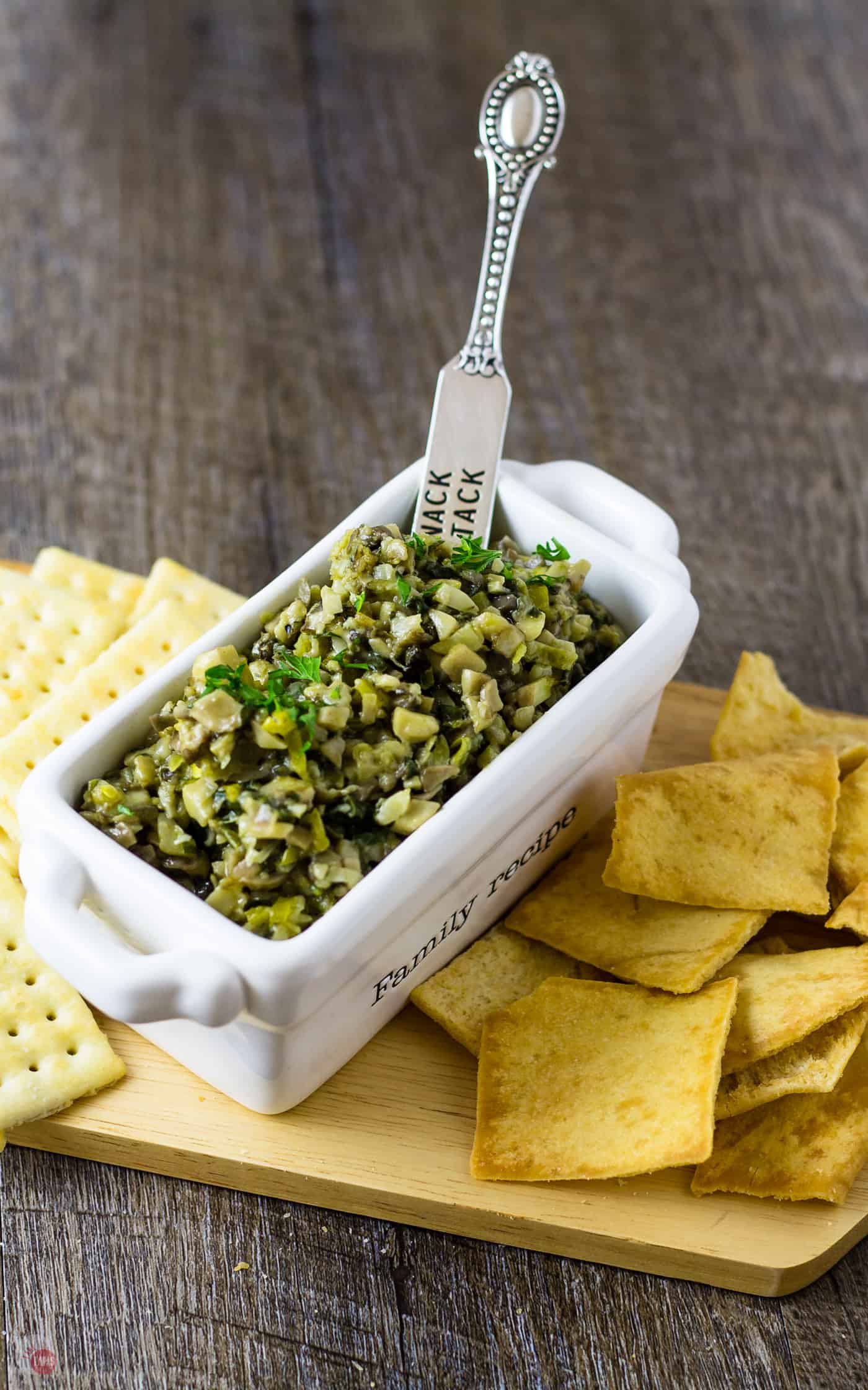 Mushroom Bruschetta spread in a white serving dish with a mini knife spreader sticking out the top and crackers sitting on a wood platter