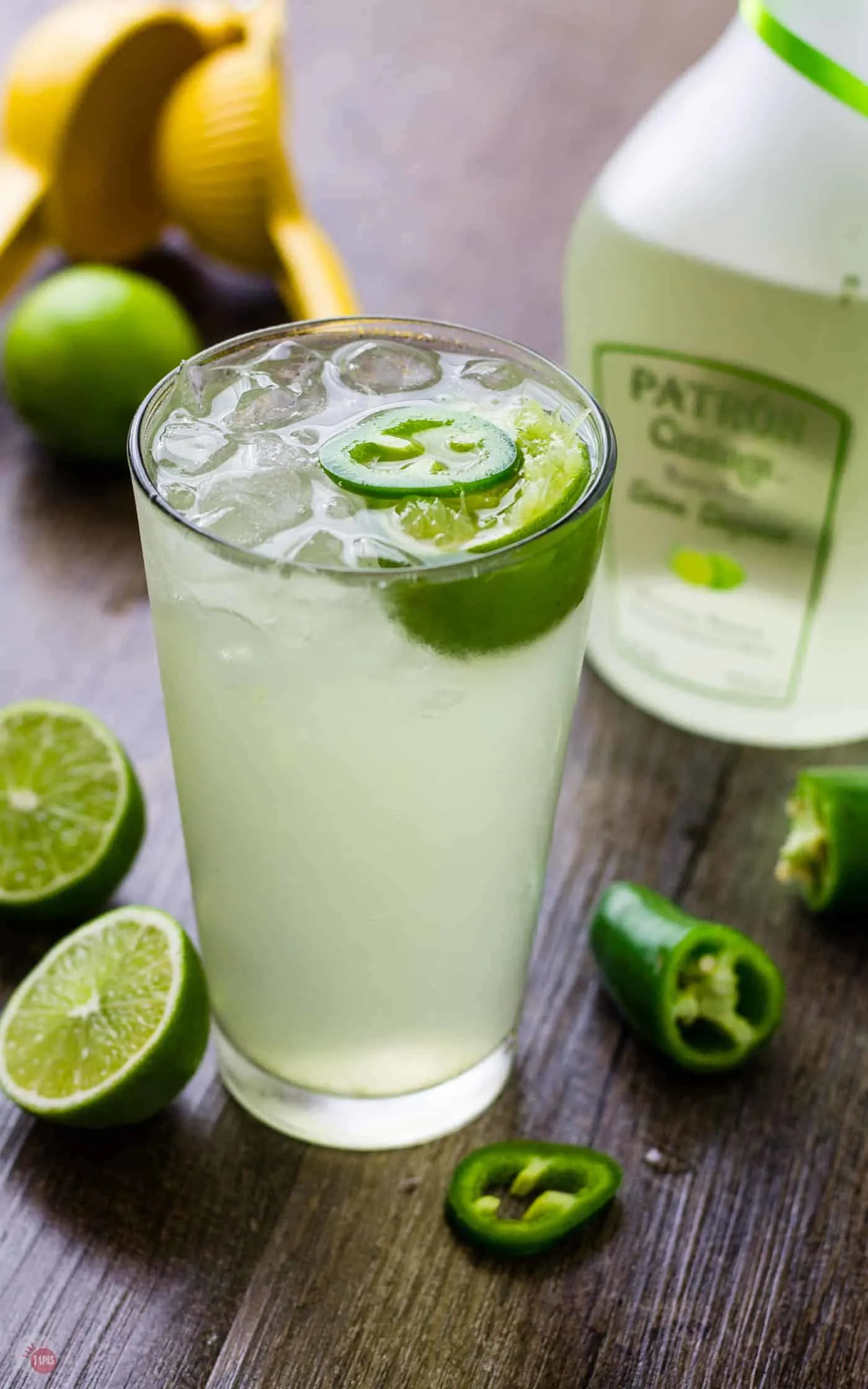 Lime Coconut Cooler Cocktail with sparkling water on a table surrounded by liquor bottle, jalapenos and cut limes.