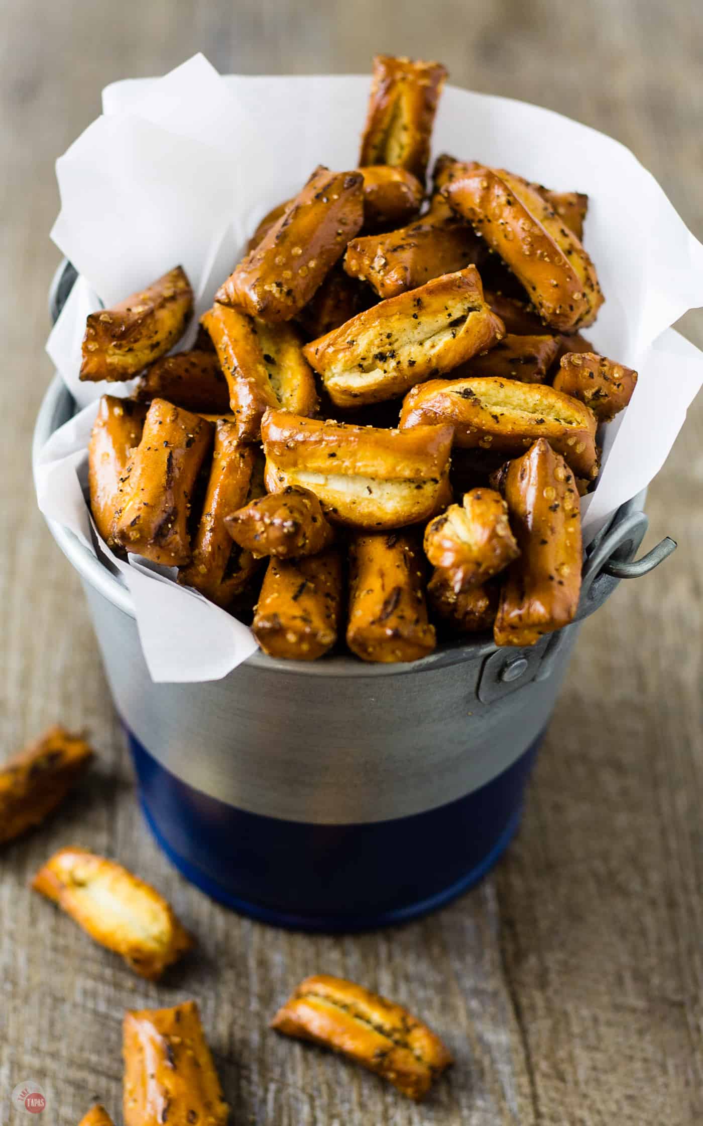 close up of Sriracha black pepper pretzels in a parchment lined metal canister with a spilled on a wood table.