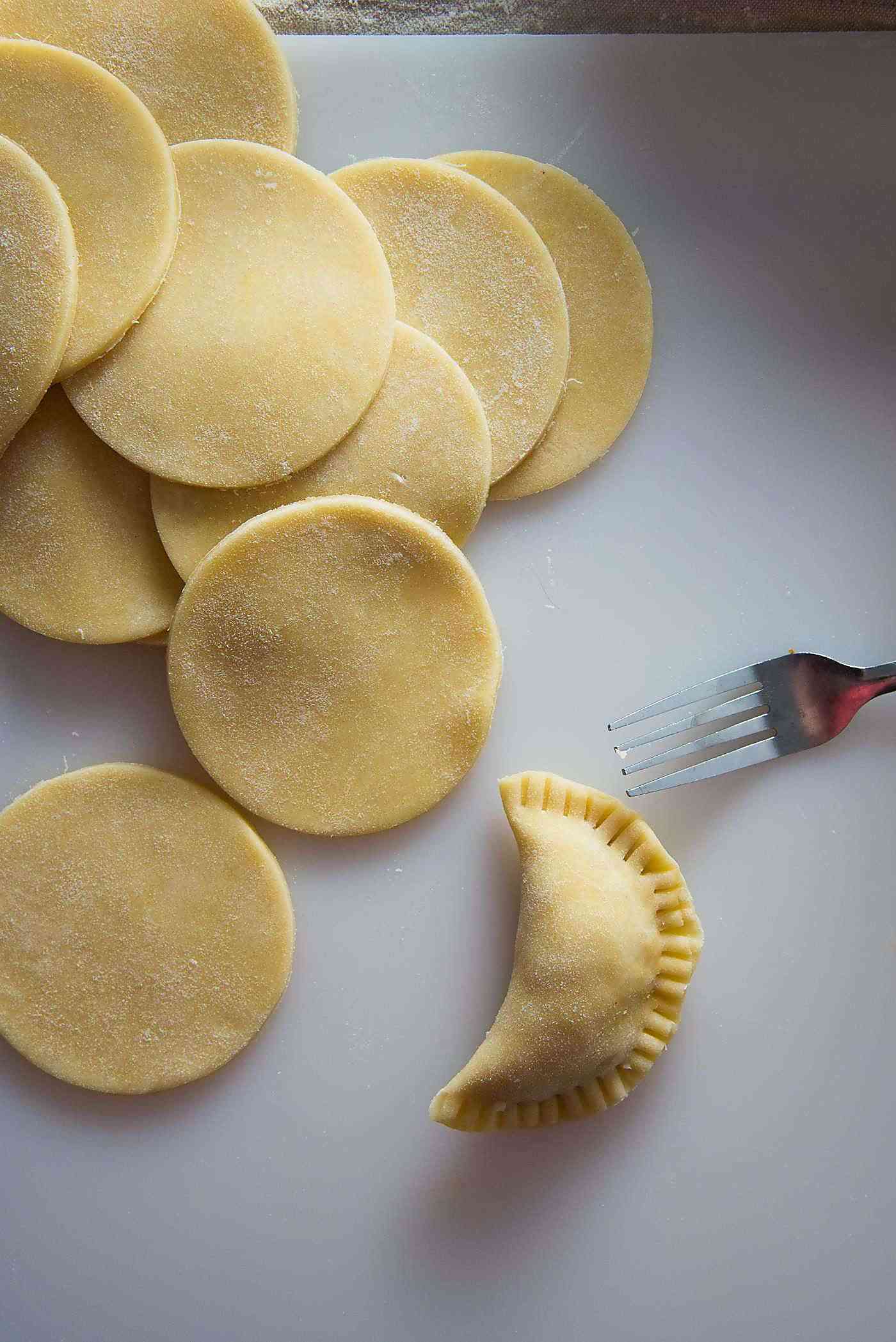 Discs of dough on the left with one empanada made, and a fork on the left, on a table.