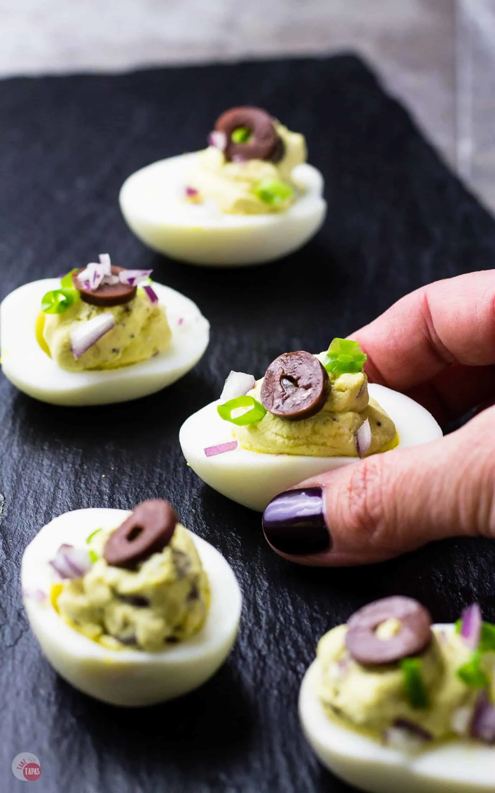 Hand holding a Mediterranean Deviled Egg with others on a black wooden platter
