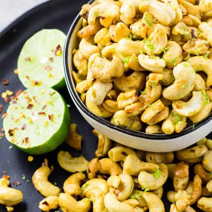 Zesty Chili Lime Cashews in a white bowl on a black plate with more cashews and a lime.