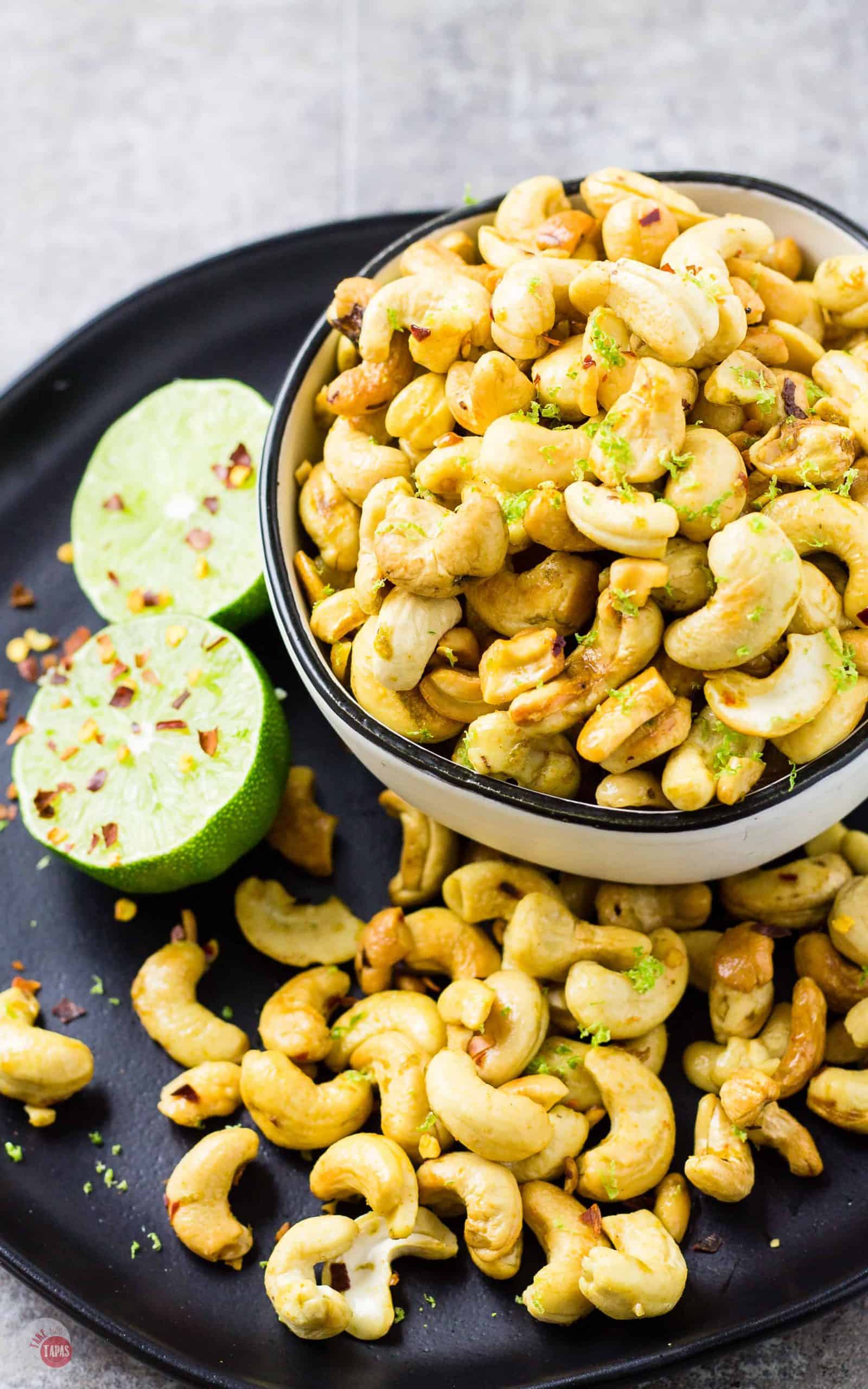 Zesty Chili Lime Cashews in a white bowl on a black plate with more cashews and a lime.