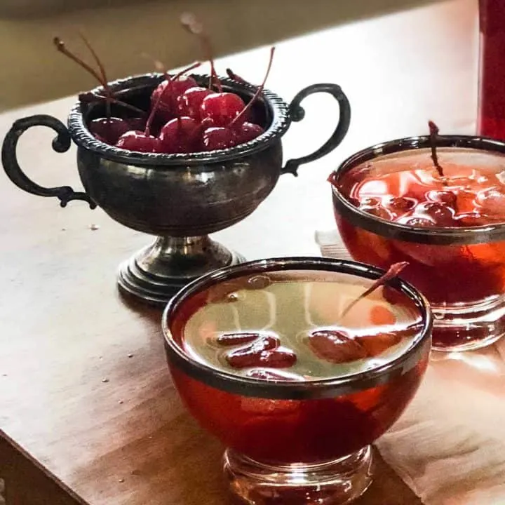 2 glasses with Cherry Manhattan punch in them and a container full of cherries on a table