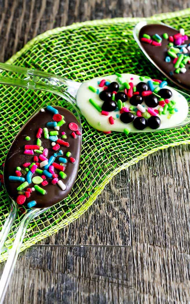 two clear spoons with chocolate and sprinkles in them on a green ribbon on a wood board