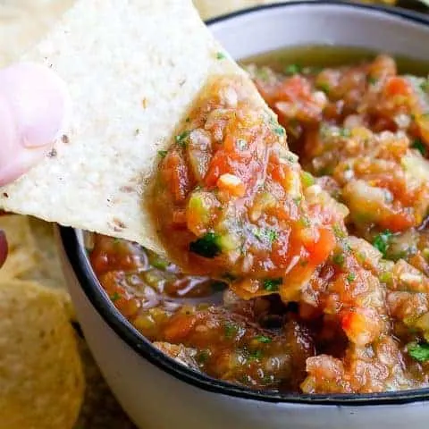 white bowl with salsa surrounded by chips with a hand scooping out salsa