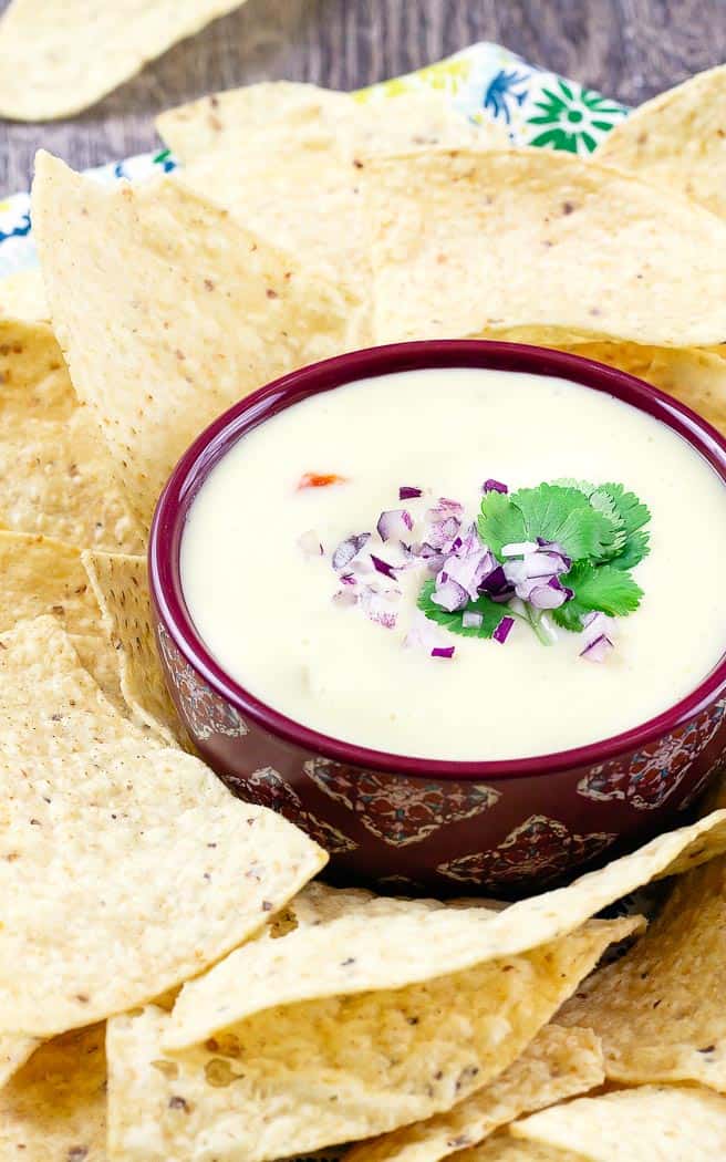 white queso dip in a red bowl topped with onions and cilantro surrounded by chips