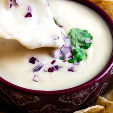 tortilla chip dipping in a red bowl of queso dip with red onion and cilantro topping