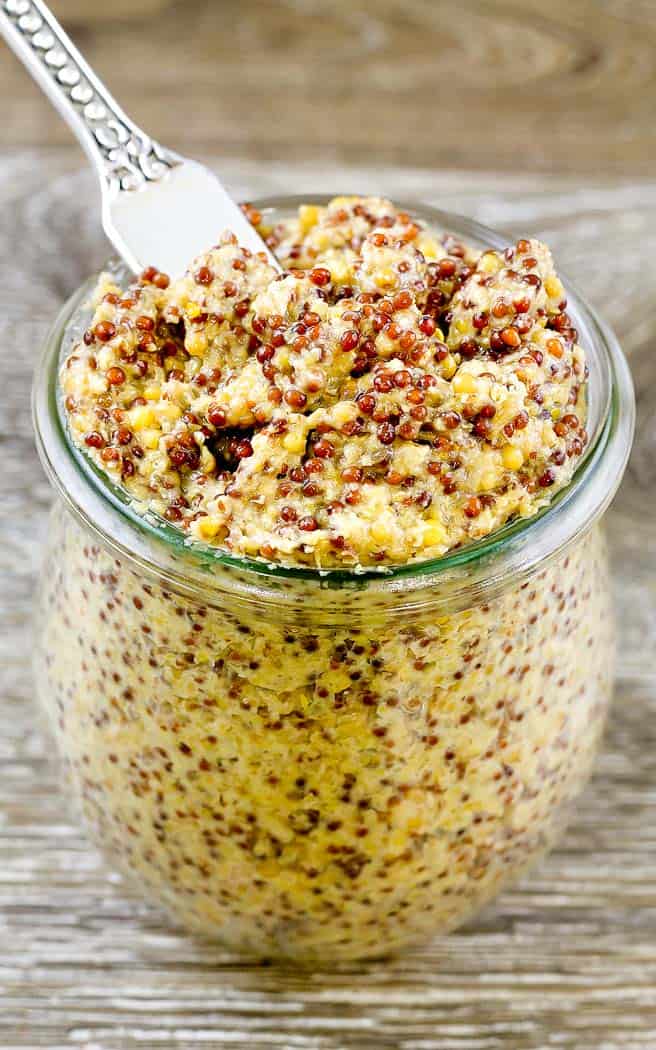 Close up of the whole grain mustard in a glass jar with a spreading knife sticking out the top