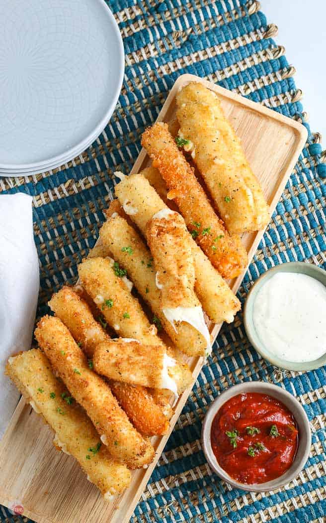 overhead picture of pile of cheese sticks with one broken in half and a small bowl of red sauce and ranch with a stack of plates