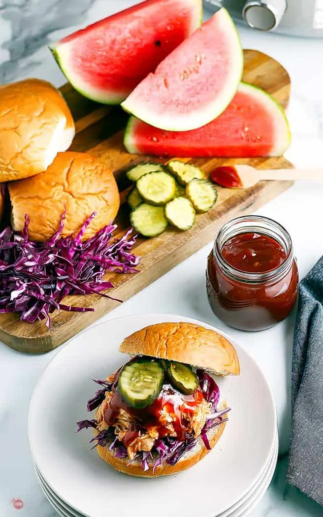 overhead picture of bbq chicken on a sandwich with pickles. Stacked on white plates with board of toppings in the background and a jar of bbq sauce