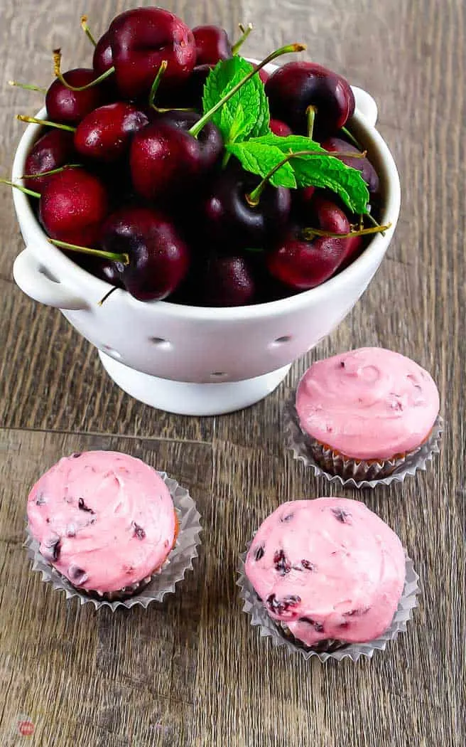 three cherry cupcakes and a bowl of cherries on a wood board