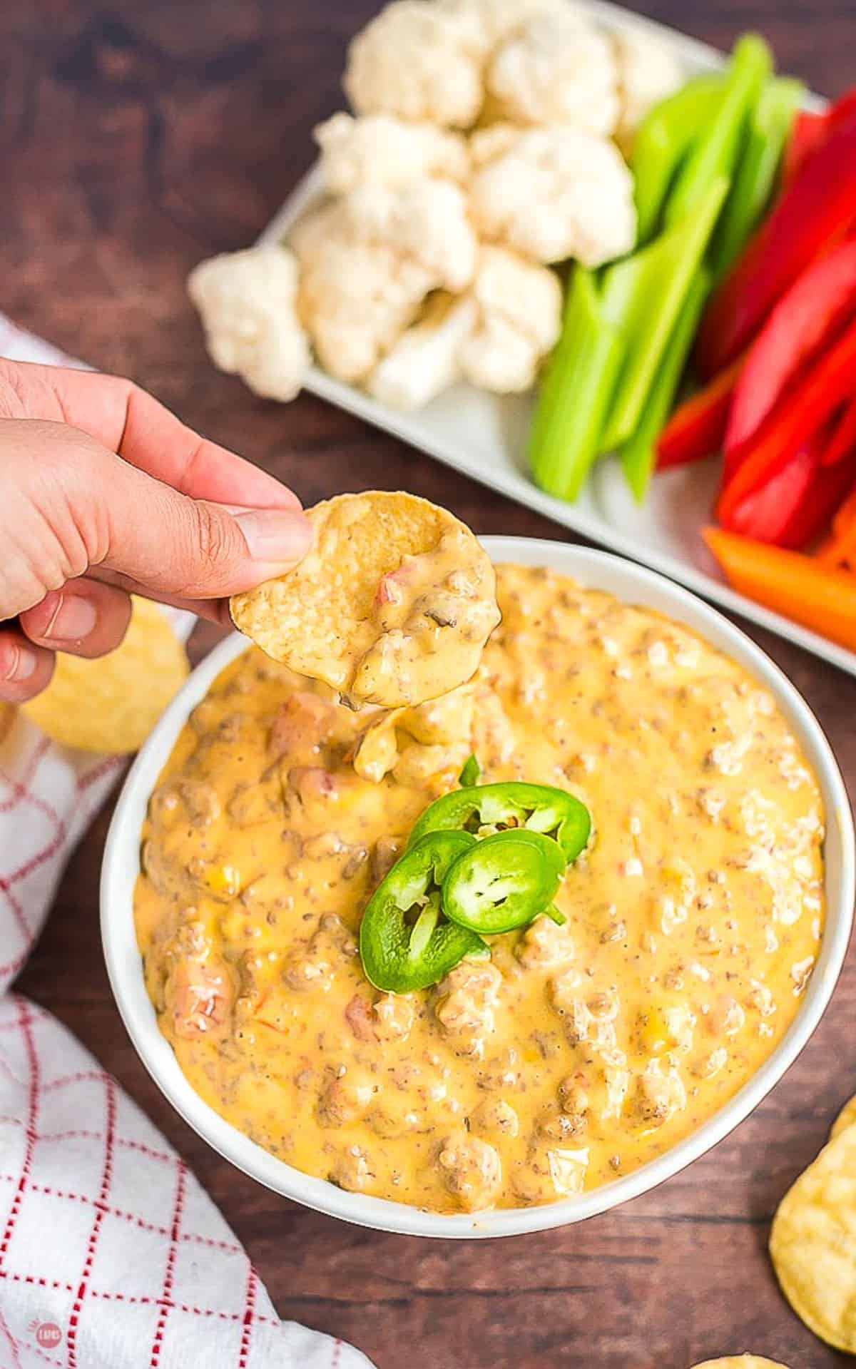 chip scooping out some cheese dip in a white bowl of cheesy beef dip topped with jalapenos with a tray of veggies in the background