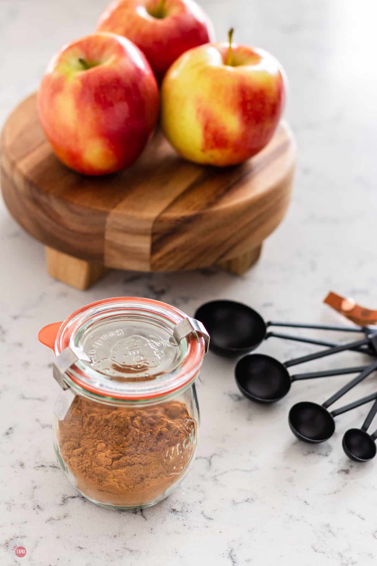 close jar of spices with a board next to it topped with apples and a set of measuring spoons