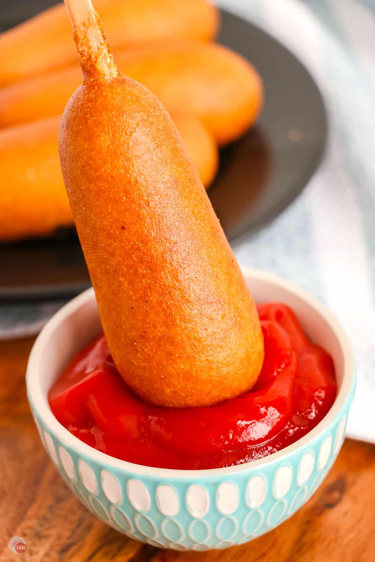 Corn Dog being dipped into a ramekin of ketchup with a black plate of corndogs in the background on a table. 