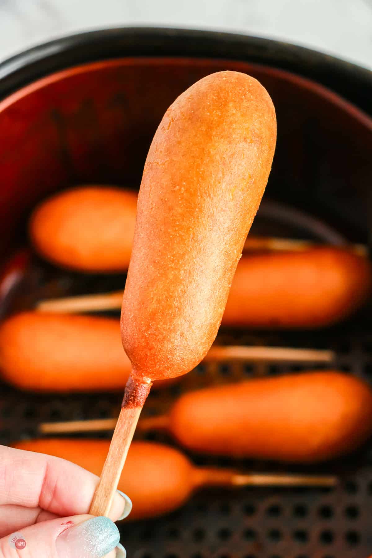 A Corn Dog is being held in mid-air over an air fryer basket full of corndogs. 
