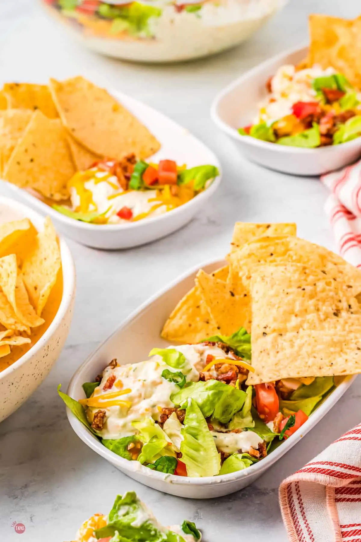 BLT Dip in a rectangle dish with chips on the side, on a table with other plates of the same thing.