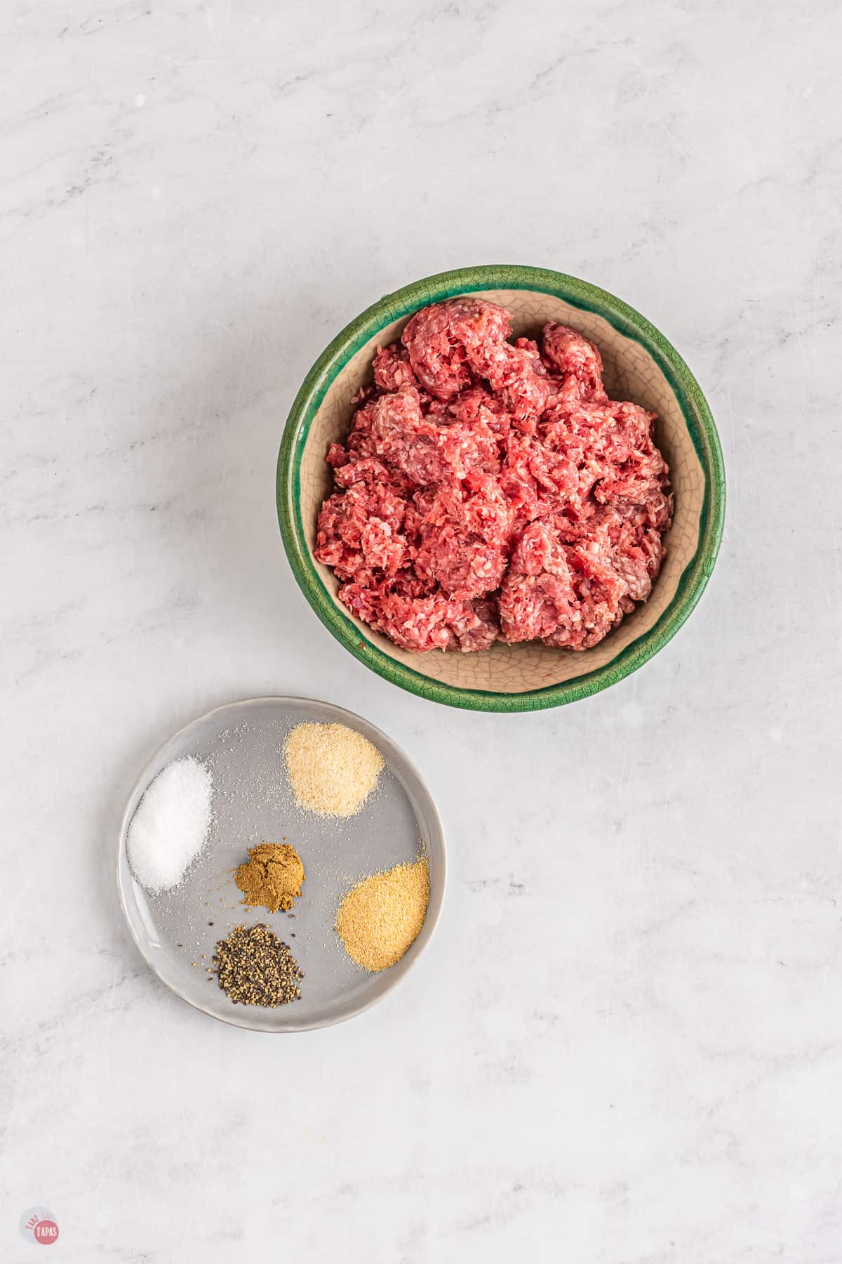 Top view of cream bowl with a green edge with raw mince in it and beside it there is a shallow, small, grey plate with salt, garlic powder, onion powder, black pepper, cumin on it. 