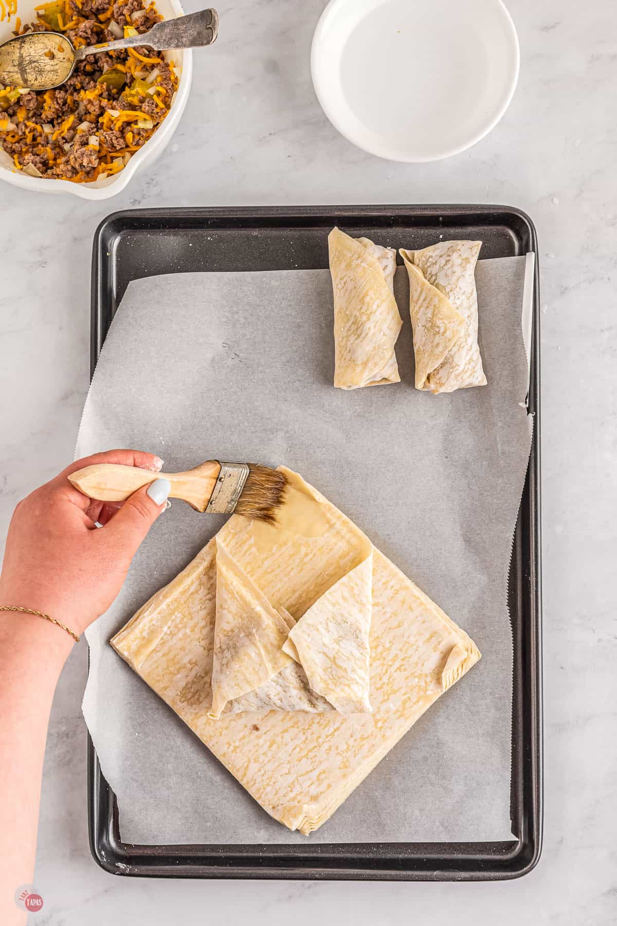 Top view of a tray lined with parchment paper with two uncooked egg rolls on it at the top of the tray and in the bottom half of the tray there is a stack of egg roll paper with the top sheet being made into an eggroll; the egg roll paper has been filled in the middle, folded with the bottom corner being folded in first, then both side corners folded in, and there is a hand brushing the top of the top corner with water. 