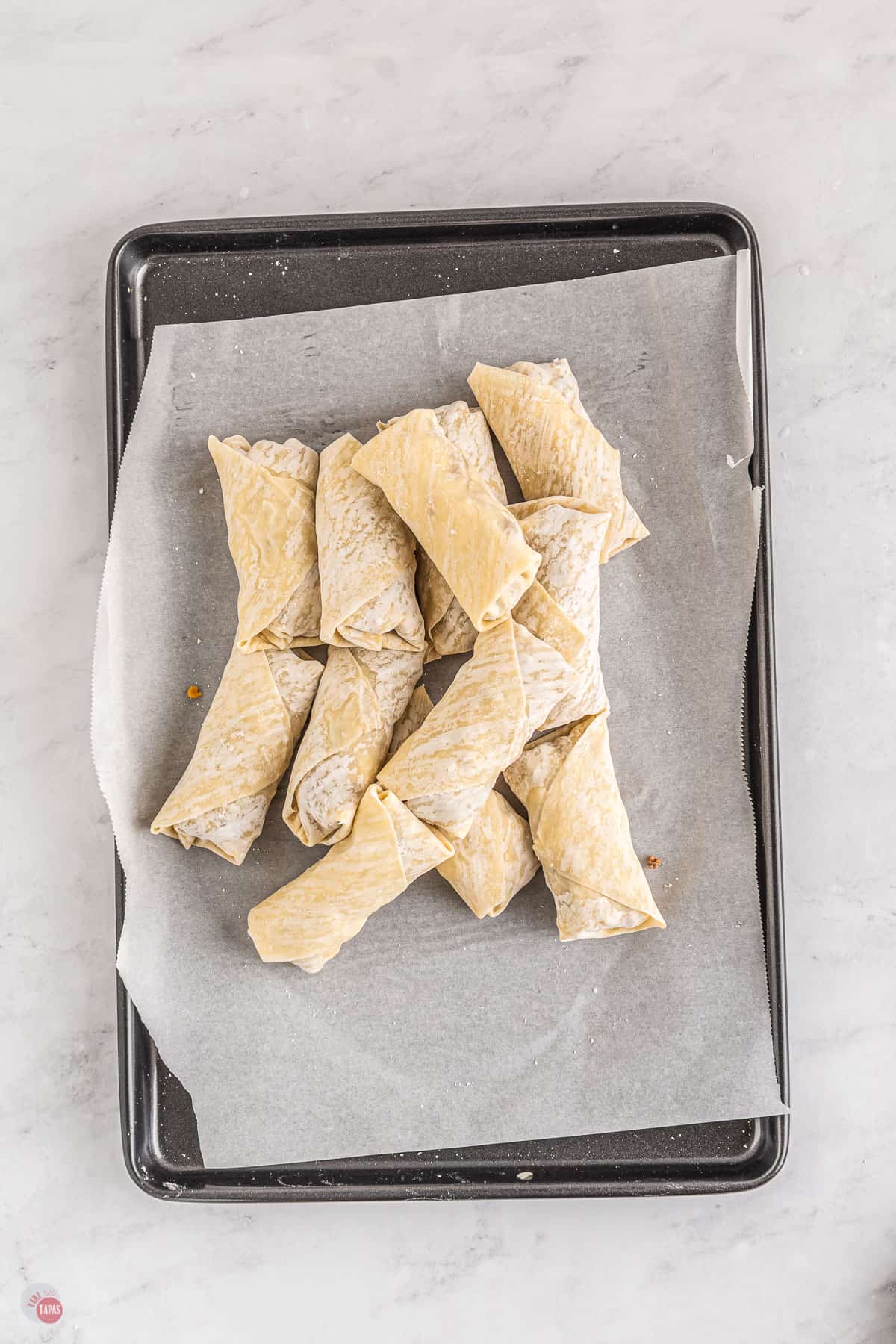 Top view of a tray lined with parchment paper with a heap of uncooked egg rolls on it. 