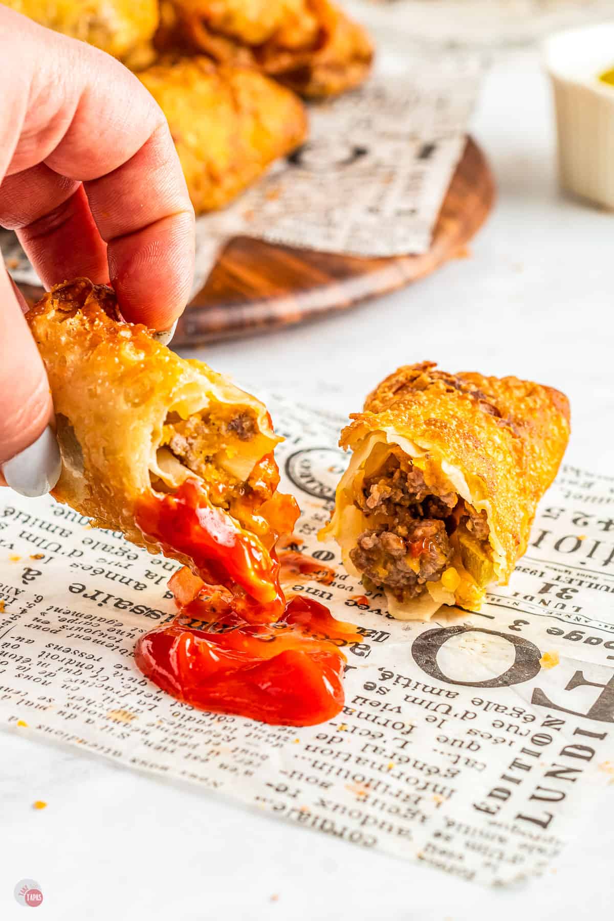 A crispy egg roll cut in half, with minced beef and cheese filling, on top of newspaper with one half being picked up and dipped into ketchup.
