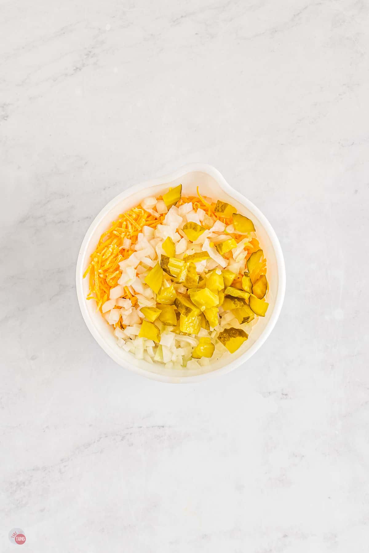 Top view of white bowl filled with diced onion, chopped pickles and shredded cheese. 