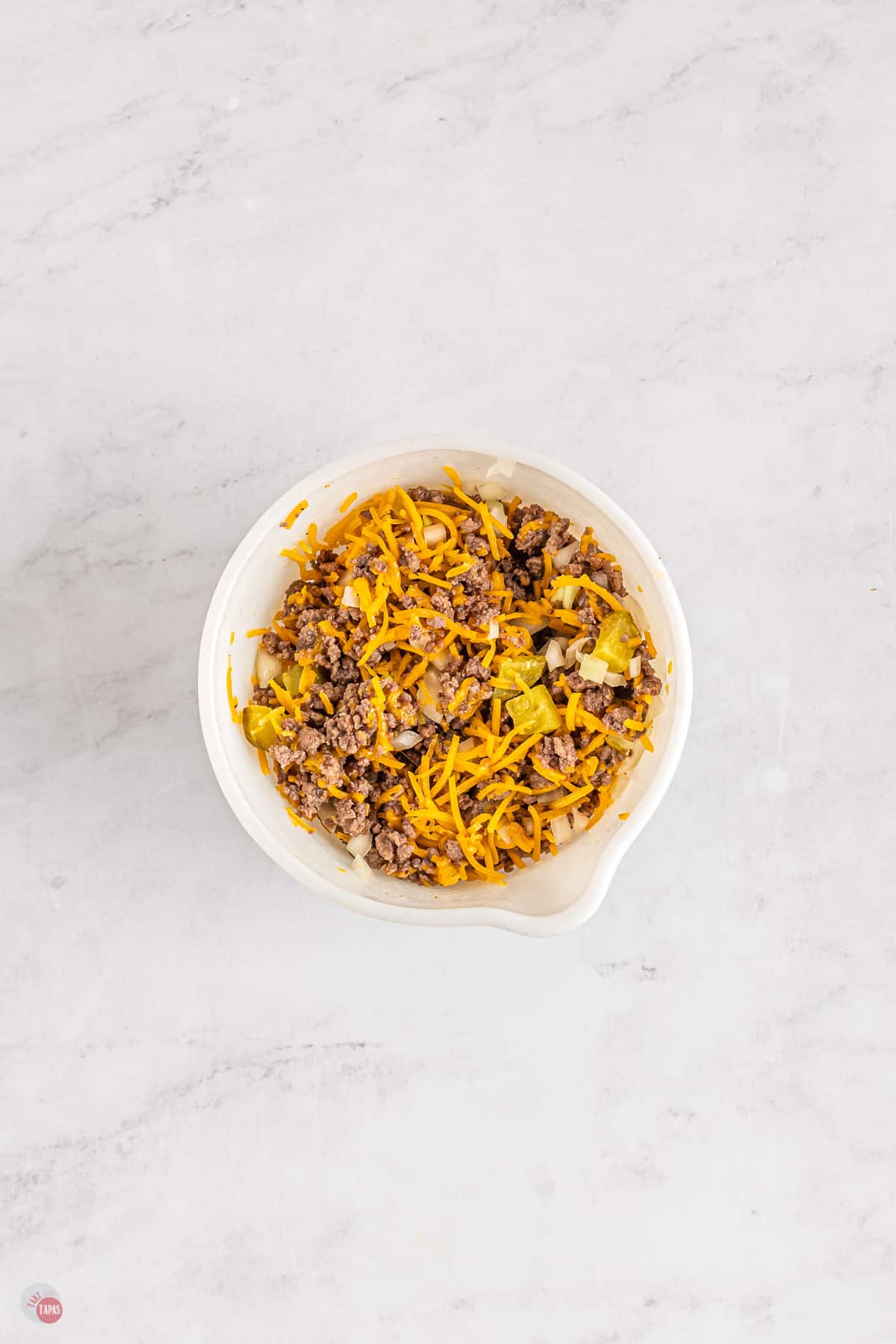 Top view of white bowl with mixture of cooked beef mince and chopped onion, diced pickles and cheese.