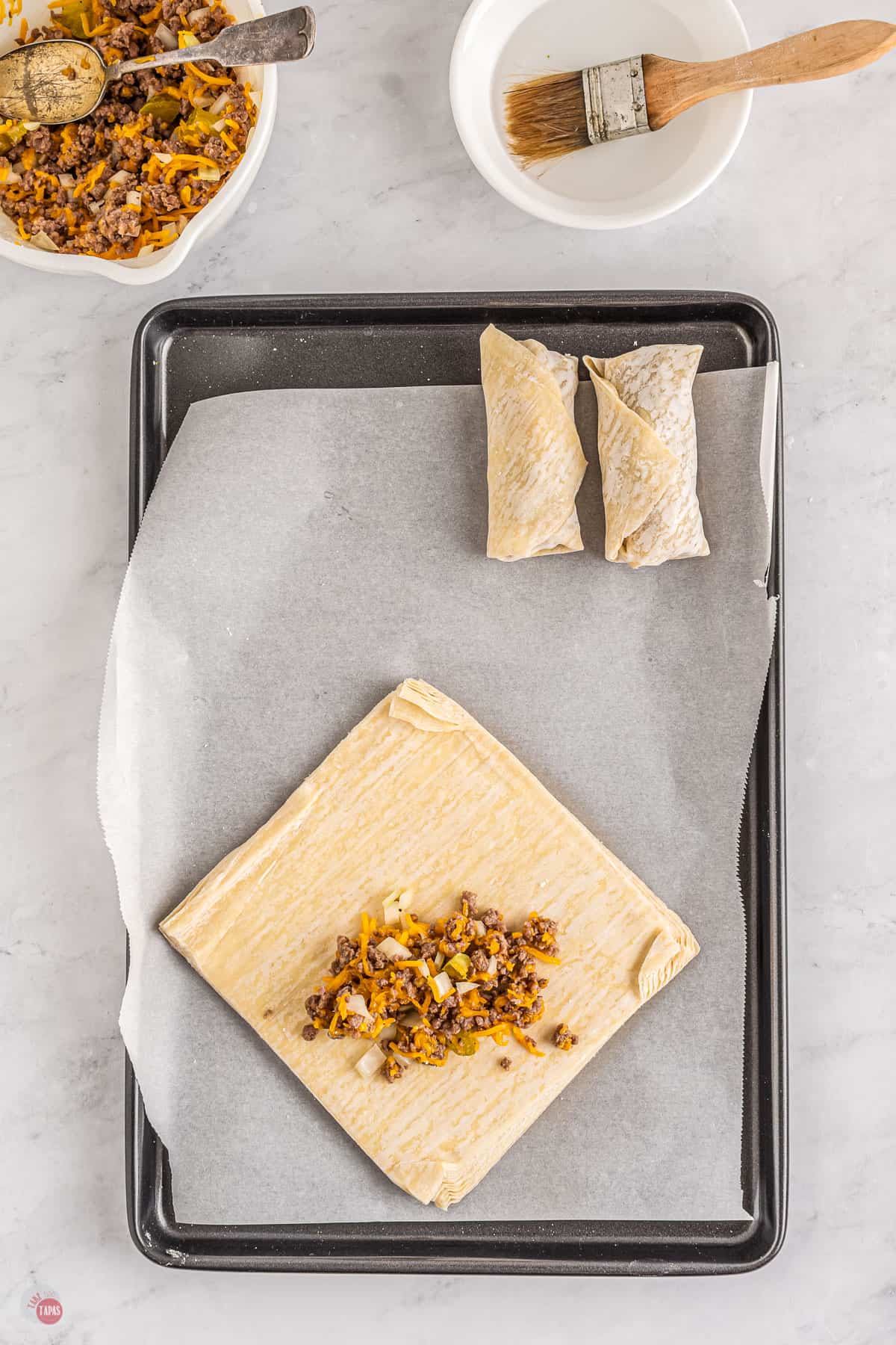 Top view of a tray lined with parchment paper with two uncooked egg rolls on it at the top of the tray and in the bottom half of the tray there is a stack of egg roll paper with the top sheet being made into an eggroll; the egg roll paper has been filled in the middle with a mixture of cooked beef mince and chopped onion, diced pickles and cheese.