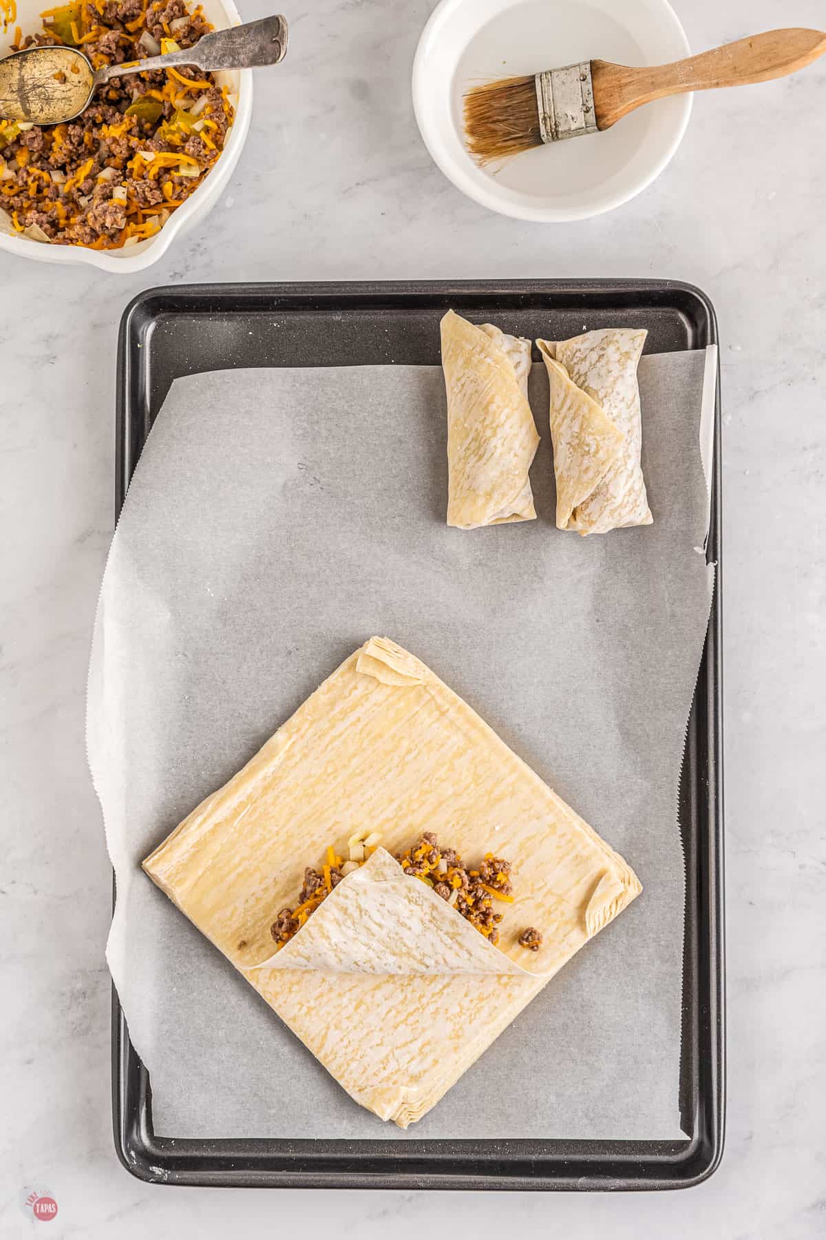 Top view of a tray lined with parchment paper with two uncooked egg rolls on it at the top of the tray and in the bottom half of the tray there is a stack of egg roll paper with the top sheet being made into an eggroll; the egg roll paper has been filled in the middle, folded with the bottom corner being folded up.