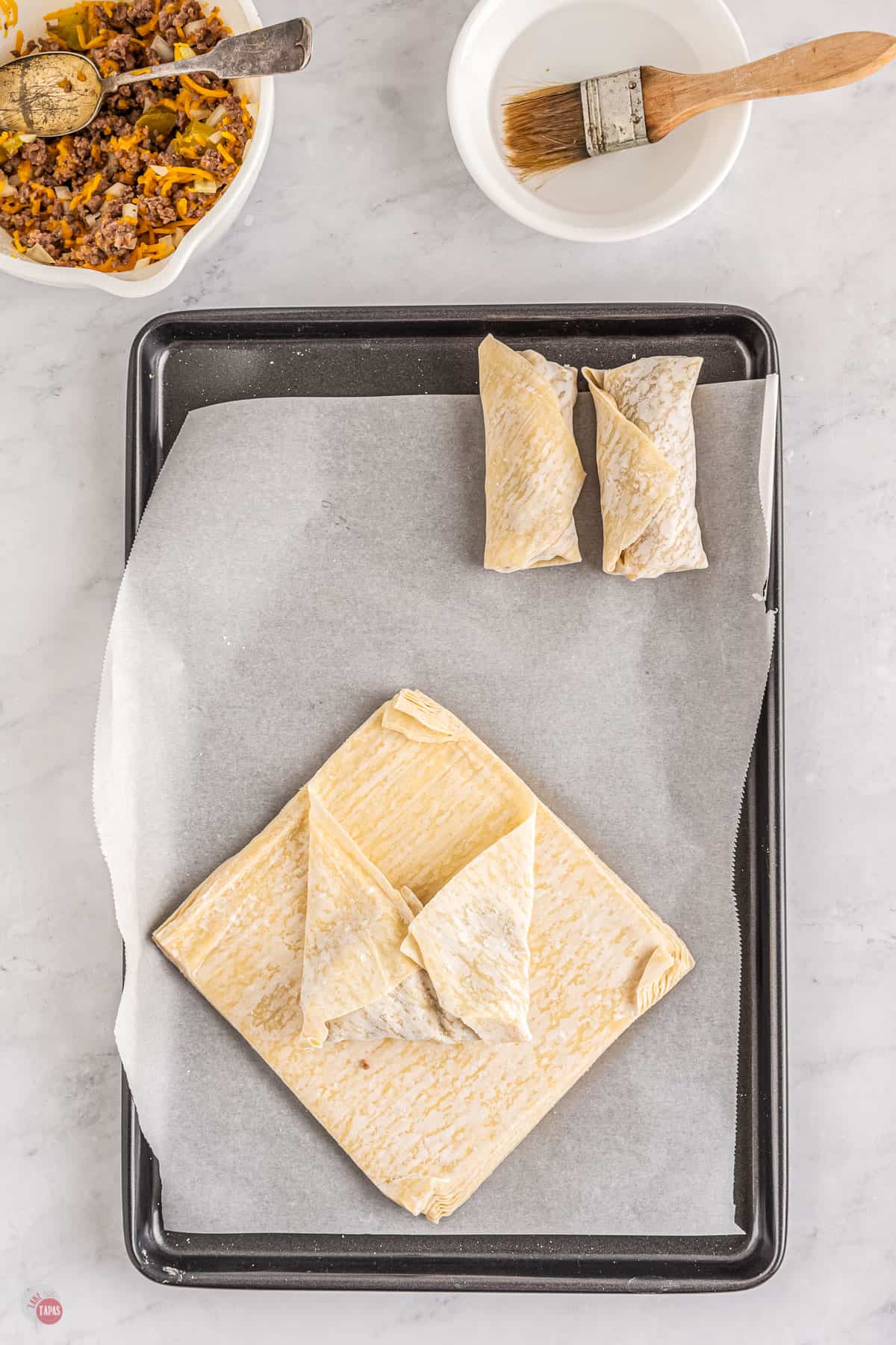 Top view of a tray lined with parchment paper with two uncooked egg rolls on it at the top of the tray and in the bottom half of the tray there is a stack of egg roll paper with the top sheet being made into an eggroll; the egg roll paper has been filled in the middle, folded with the bottom corner being folded in first, then both side corners folded in.