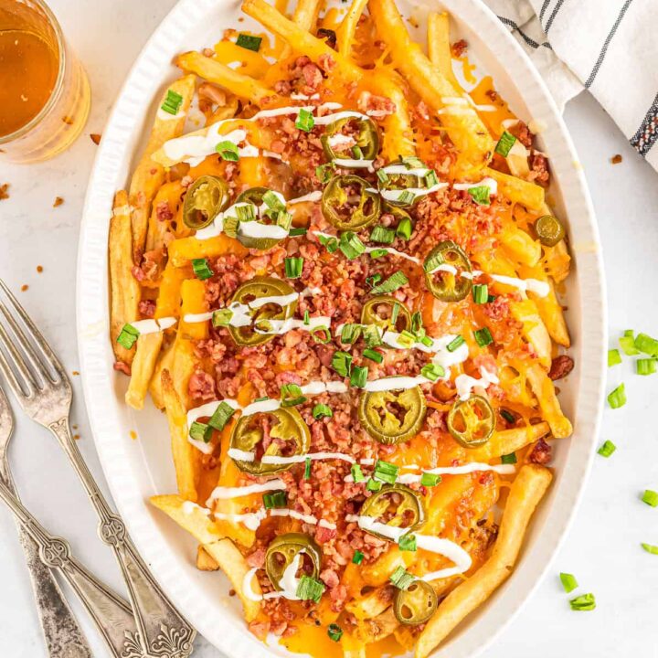French fries loaded with cheese, bacon, and jalapeños, topped with green onions and drizzled with sour cream in a white baking dish on a table.