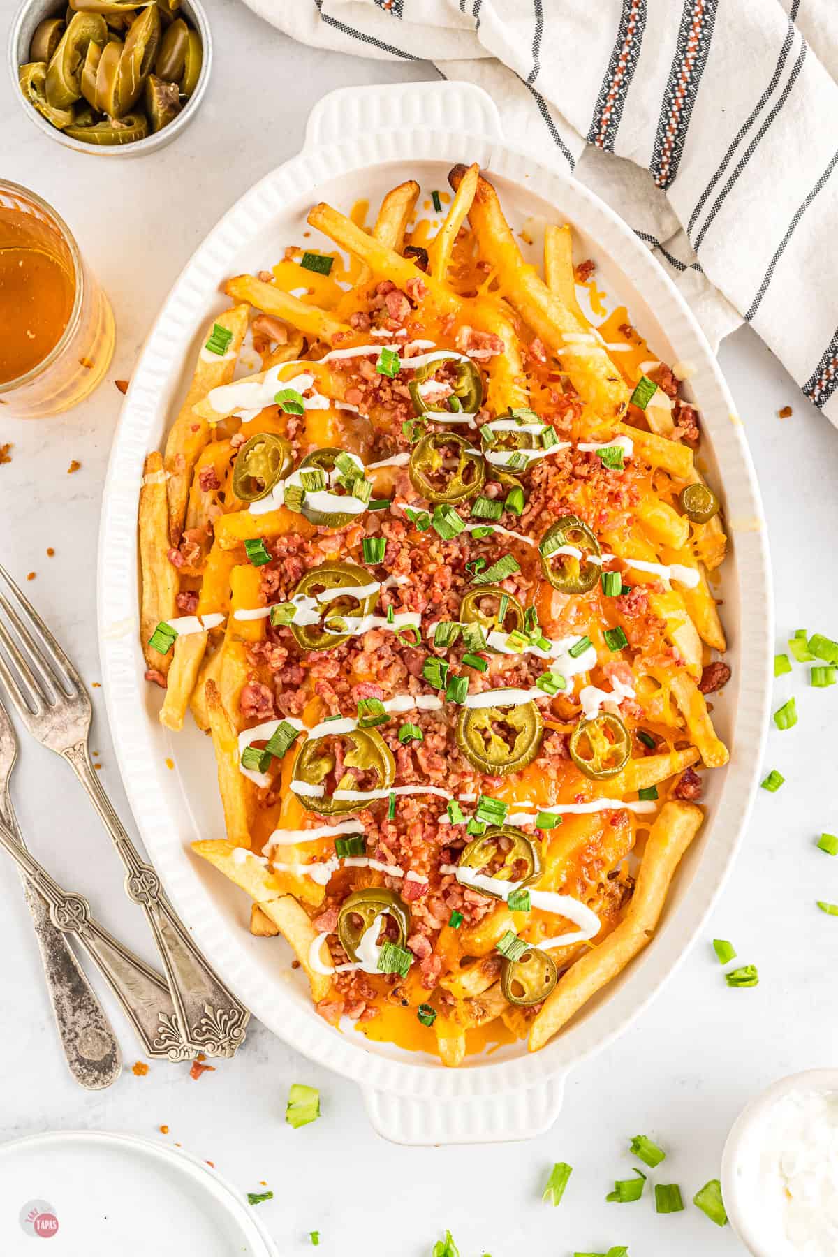 French fries loaded with cheese, bacon, and jalapeños, topped with green onions and drizzled with sour cream in a white baking dish on a table.