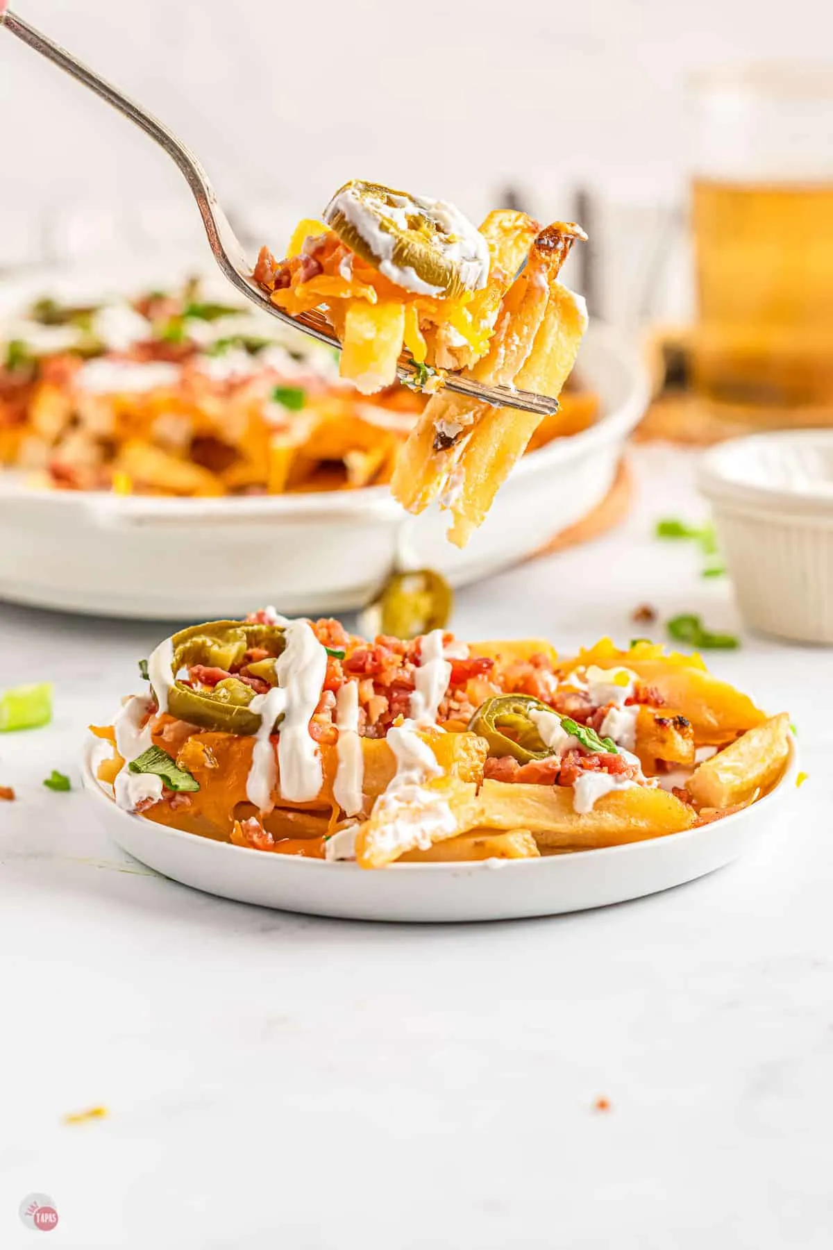 A forkful of loaded fries is being lifted mid-air above a plate full of loaded fries with a casserole dish of bacon and jalapeño loaded fries in the background. 